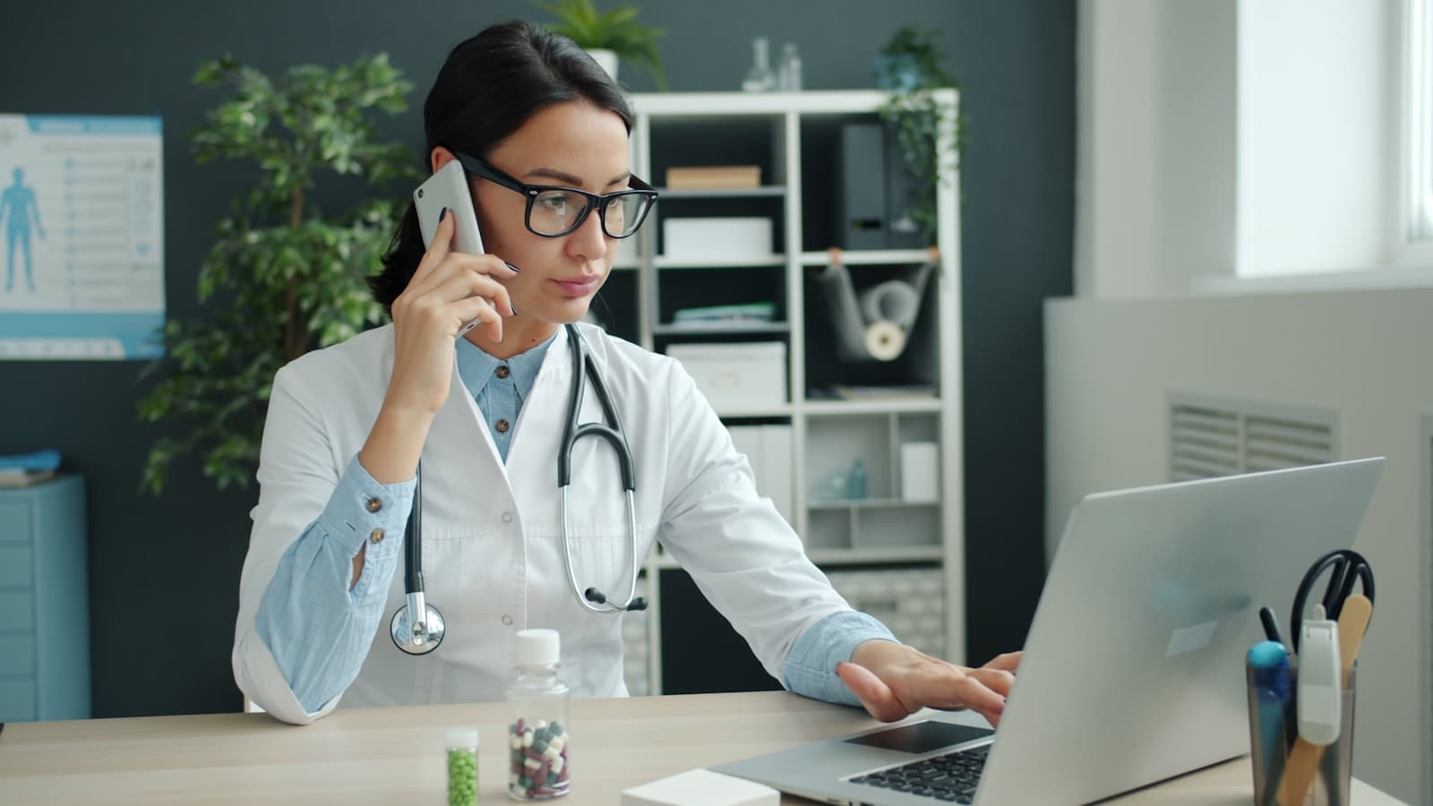 Médica atende ao telefone enquanto consulta a agenda no laptop do consultório